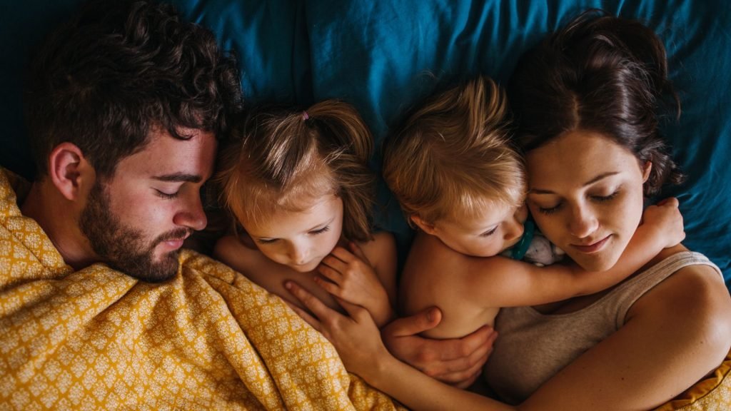 Clocks go back this weekend — the top 3 problems parents will face with their kids sleep and how to fix them fast A family sleeps in bed together, though the two children are both awake.