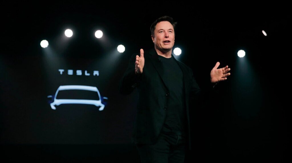 Elon Musk on stage at an event with the word 'Tesla' displayed behind him