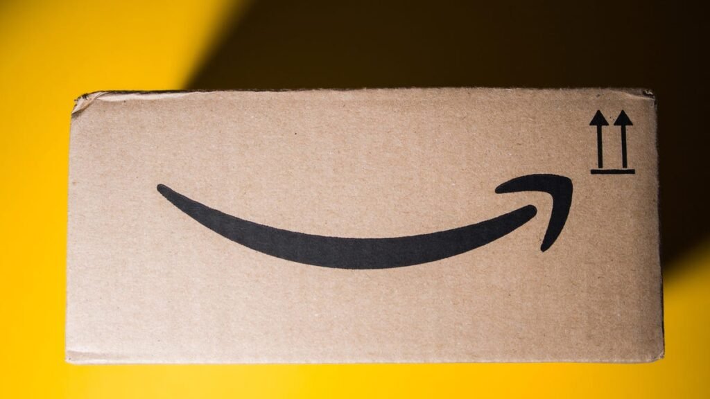 A side view of an Amazon delivery box rests against a bright yellow background. The top right corner is covered in shade from the box's shadow.