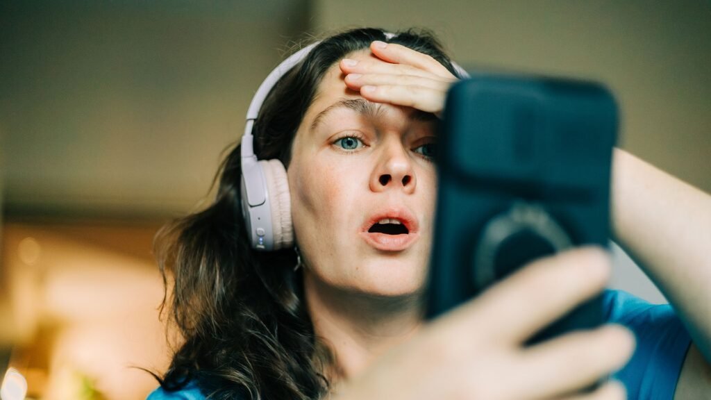 A woman in headphones looks shocked at her phone, holding her hand to her head