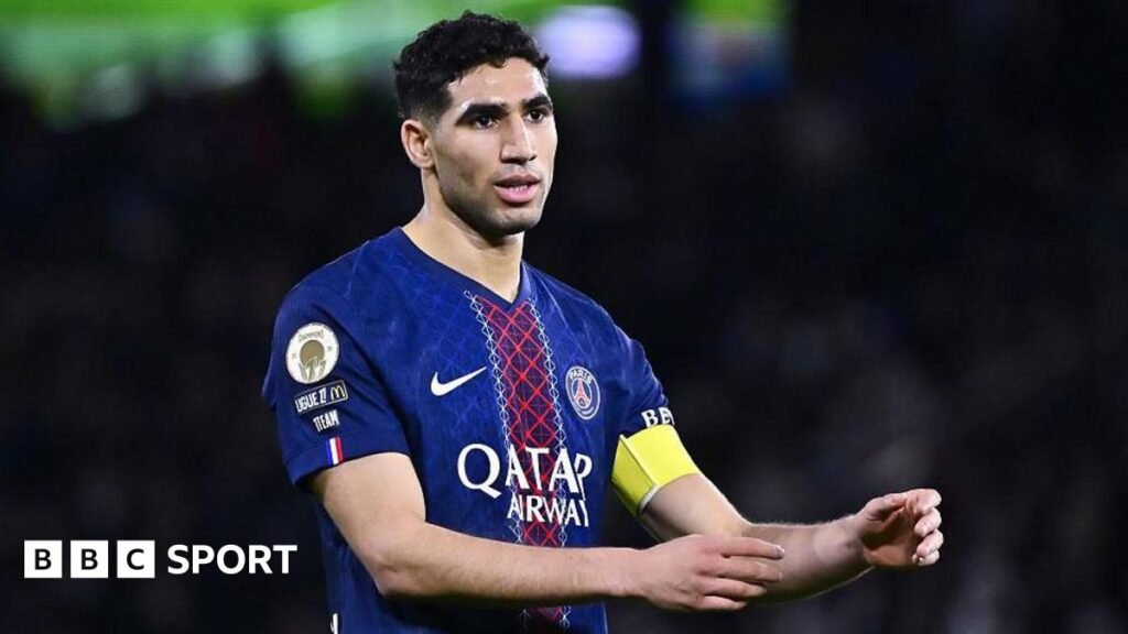 Achraf Hakimi, wearing a blue and red Paris St-Germain shirt, holds out his arms