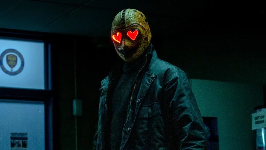A person in all black, wears a mask with red hearts for eyes.