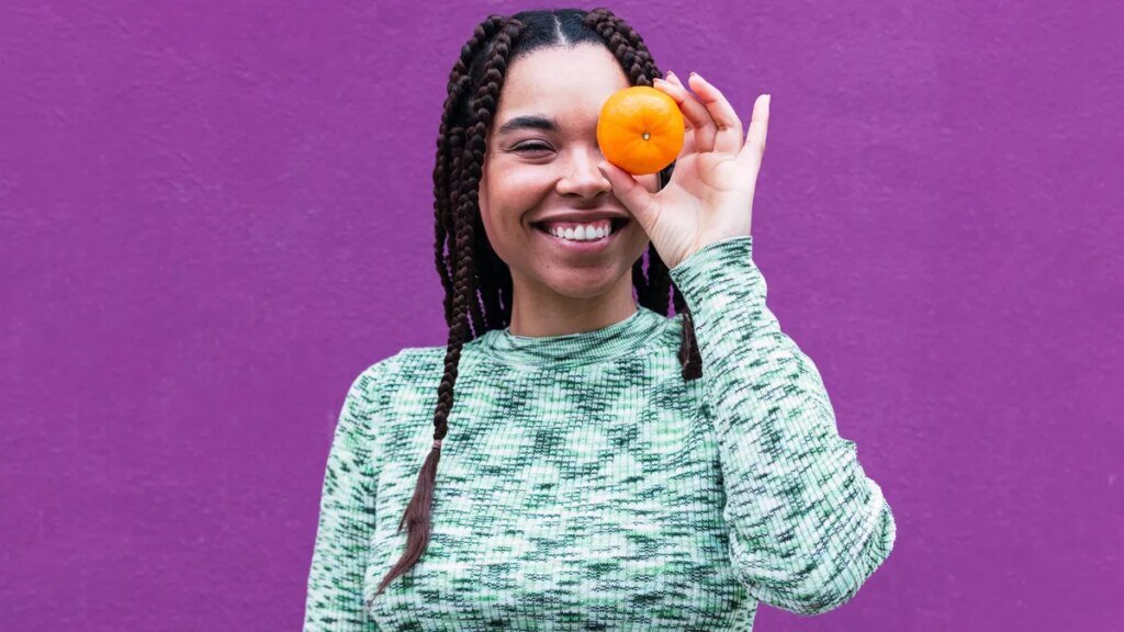 These Superfoods Will Keep Your Vision Sharp as You Age A person with long brown braided hair and a teal patterned shirt holding an orange up in front of their left eye.